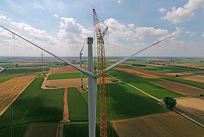 Drohnenaufnahme eines Windrads in der landwirtschaftlich genutzten Landschaft, bei dem ein großer Gittermastkran neben dem Turm steht und die Rotorblätter während der Montage weit über die grün-braun karierten Felder hinausragen.