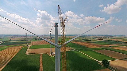 Windenergieanlage im Bürgerwindpark Adelhofen-Gollachostheim während der Rotorblattmontage – umgeben von weiteren Anlagen, Feldern und ländlicher Infrastruktur unter wolkigem Himmel.