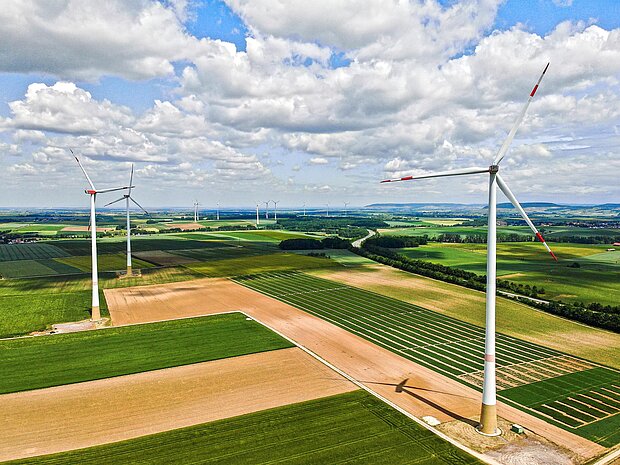 Drohnenaufnahme einer weiten Agrarlandschaft mit mehreren Windrädern, im Vordergrund ein besonders hohes Windrad über rechteckig gegliederten Feldern, unter einem Himmel mit vielen weißen Wolken.