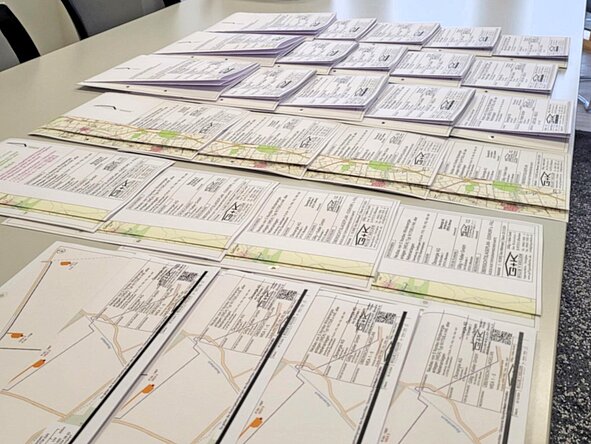 Mehrere Reihen ausgedruckter Lagepläne und Antragsunterlagen liegen ordentlich sortiert auf einem langen Konferenztisch in einem Besprechungsraum mit großen Fenstern und Büropflanzen im Hintergrund.