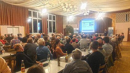 Teilnehmer der Bürgerinfo zum Bürgerwindpark Auernhofen verfolgen eine Präsentation in einem festlich dekorierten Saal mit Tischen, Getränken und Projektionsleinwand.