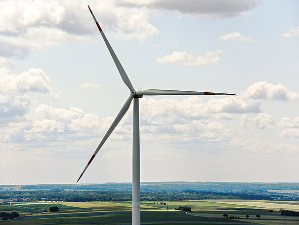 Nahaufnahme eines einzelnen Windrads vor leicht bewölktem Himmel, im Hintergrund eine weite, grüne Hügellandschaft mit Feldern und Waldstreifen.