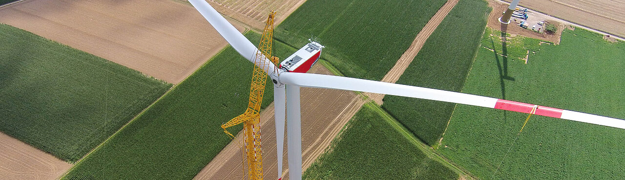 Großer gelber Kran hebt das dritte Rotorblatt zur Montage an einer Windenergieanlage im Bürgerwindpark Adelhofen-Gollachostheim – umgeben von Feldern, Baugeräten und ländlicher Infrastruktur.