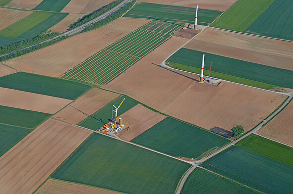 Luftaufnahme einer landwirtschaftlich genutzten, in rechteckige Felder gegliederten Landschaft, in der mehrere Windkraftanlagen im Bau sind – zu sehen sind bereits errichtete Türme und eine Baustelle mit Kran und Baumaschinen.
