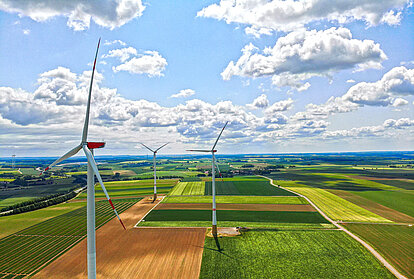 Luftaufnahme von drei Windrädern, die sich über einer weitläufigen Agrarlandschaft erstrecken. Die Felder sind in verschiedenen Grüntönen und Erdbraun unterteilt, während weiße Wolken über dem blauen Himmel schweben.