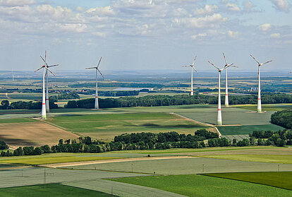 Panoramaaufnahme eines Windparks in einer weiten, leicht hügeligen Agrarlandschaft, in der mehrere hohe Windräder zwischen Feldern, Waldstreifen und kleinen Ortschaften in den Himmel ragen.