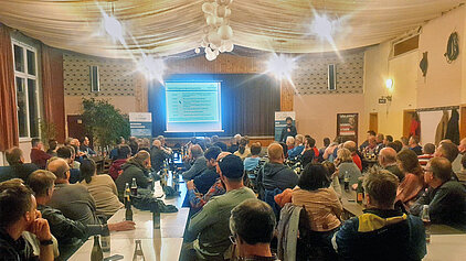 Teilnehmer der Bürgerinfo zum Bürgerwindpark Auernhofen verfolgen eine Präsentation in einem festlich dekorierten Saal mit Tischen, Getränken und Projektionsleinwand.